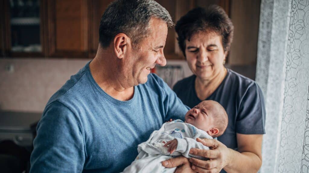 Smiling older couple lovingly holding a newborn baby in a cozy, sunlit kitchen.