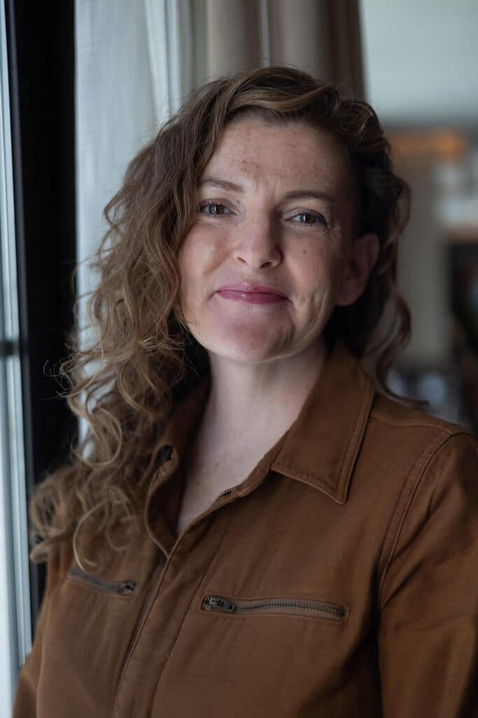 Woman with wavy brown hair in a brown zip-up top, smiling softly by a window with natural light.