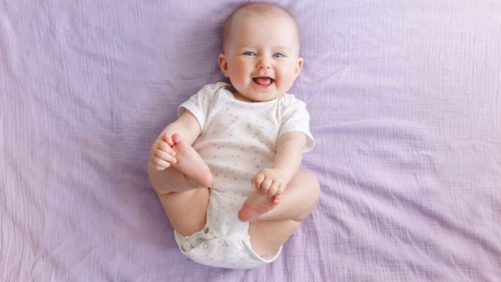 Smiling baby in a white onesie lying on a light purple blanket, holding its feet.