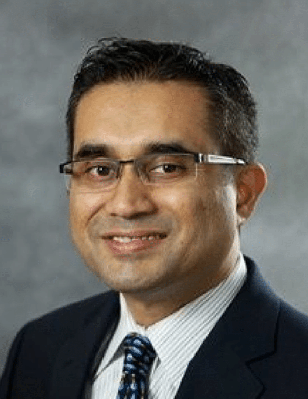 Smiling man in a suit and glasses with short dark hair, posing in front of a gray background.
