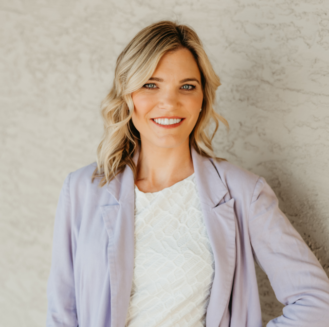 Smiling woman with blonde hair in a light purple blazer and white top, standing against a textured wall.