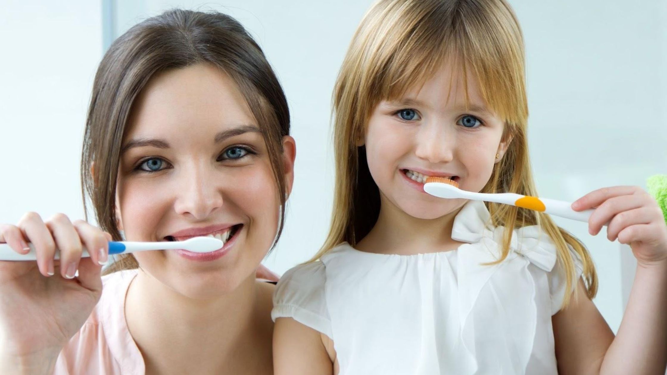 A woman and a young girl smile while brushing their teeth together with toothbrushes.