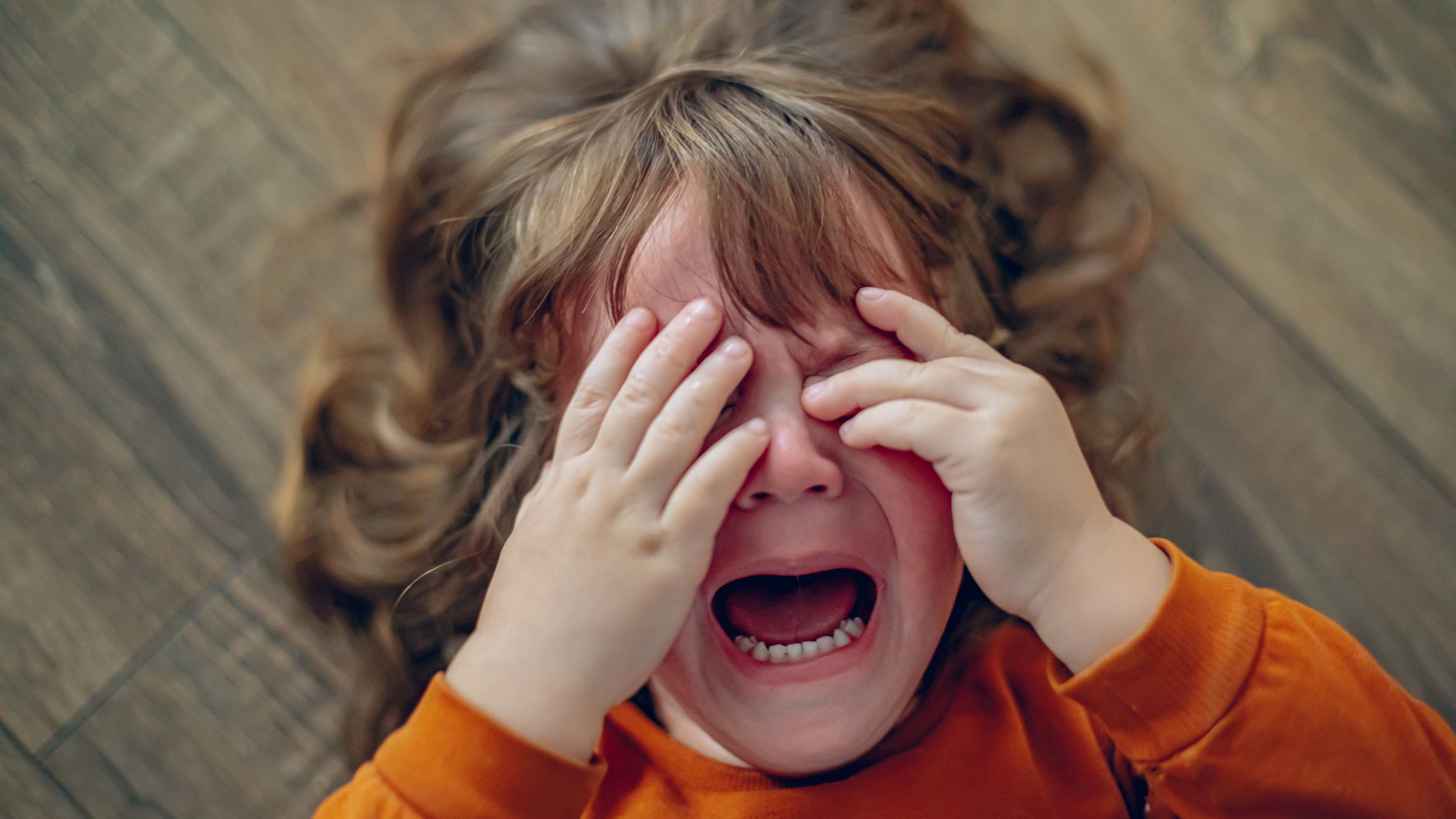 A young child lying on the floor, crying with hands covering their face.