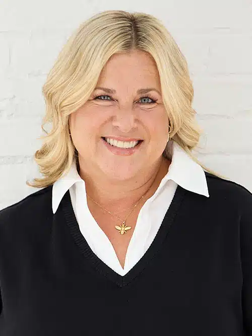 Smiling woman with blonde hair in a black sweater and white blouse, standing against a white brick wall.