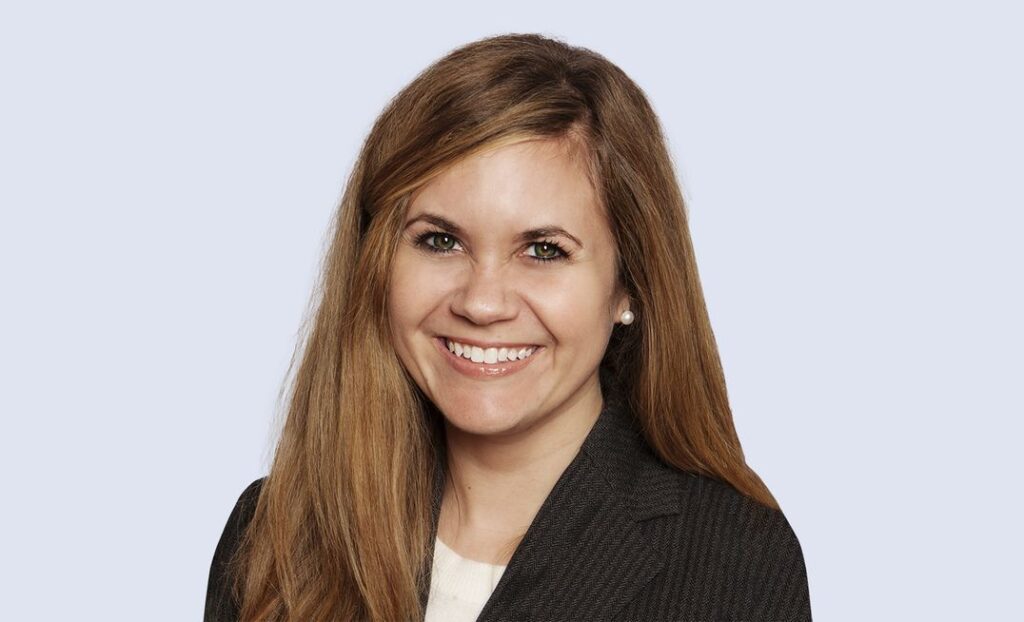 Smiling woman with long brown hair in a dark blazer, posing against a plain light background.