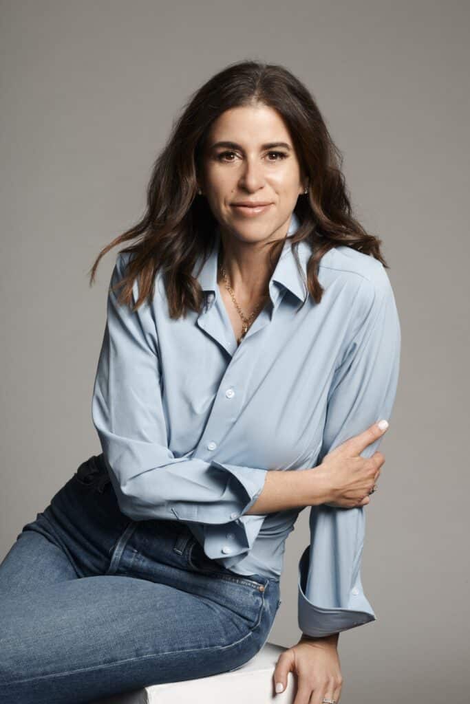 Woman with long brown hair wearing a light blue shirt and jeans, sitting and smiling at the camera.
