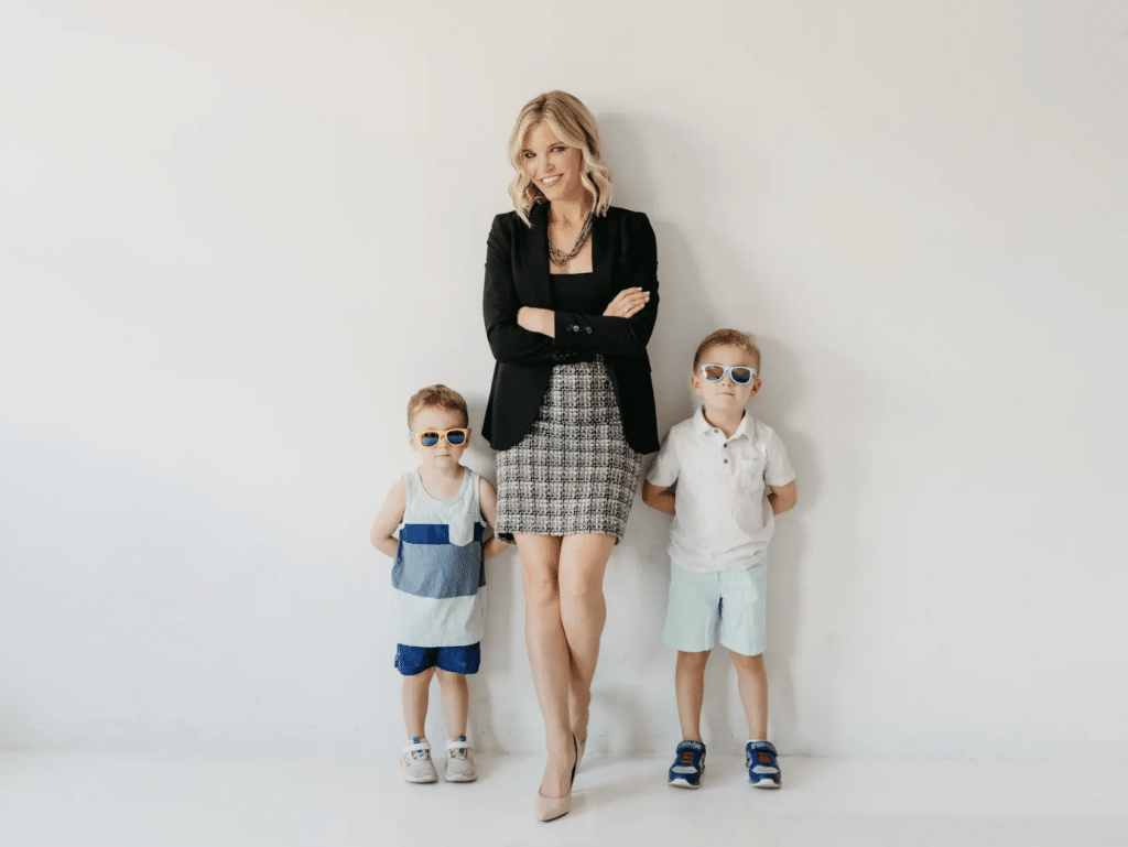 A woman in business attire stands smiling between two young boys wearing sunglasses against a white wall.