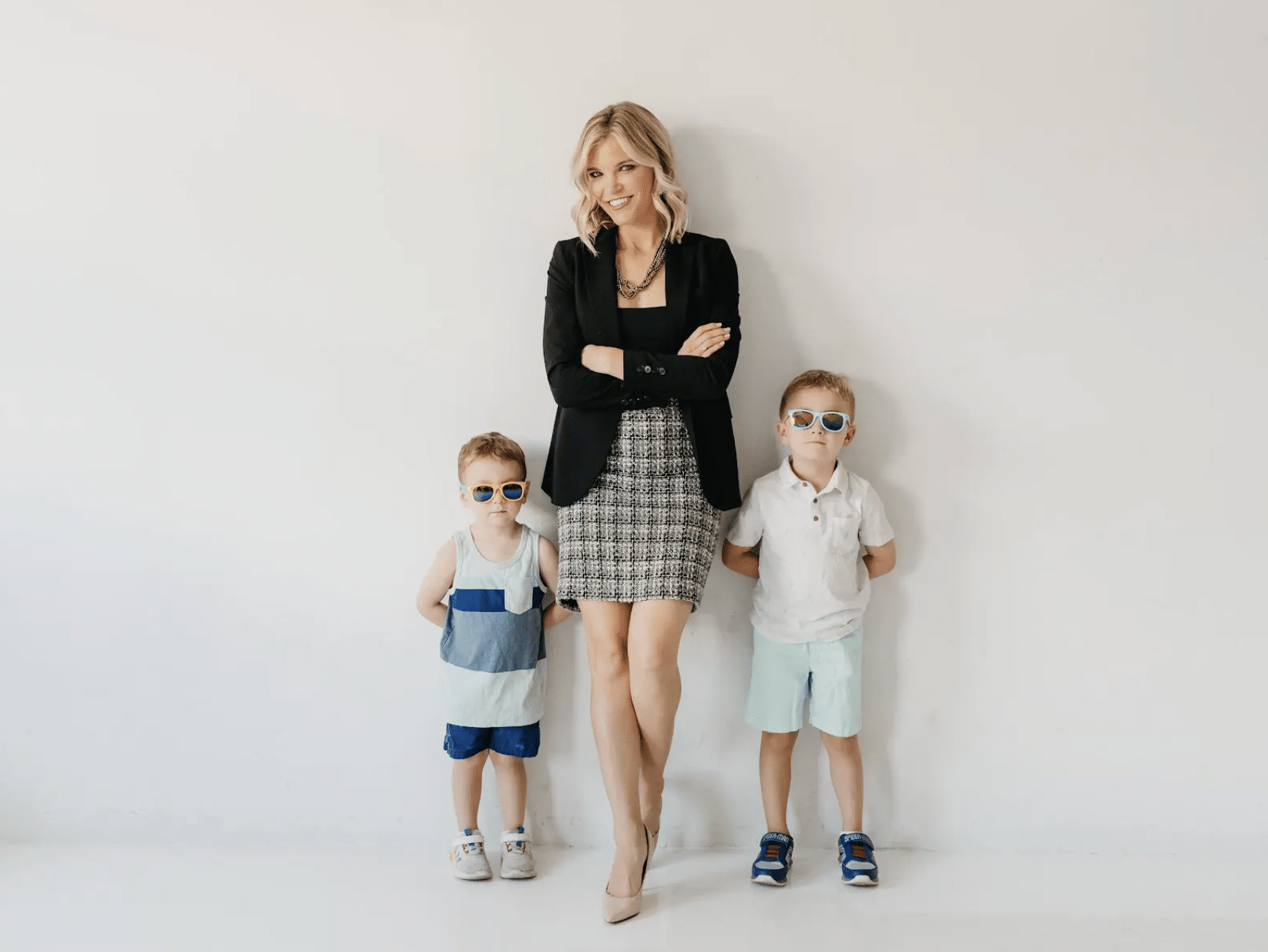 A woman in business attire stands smiling between two young boys wearing sunglasses against a white wall.