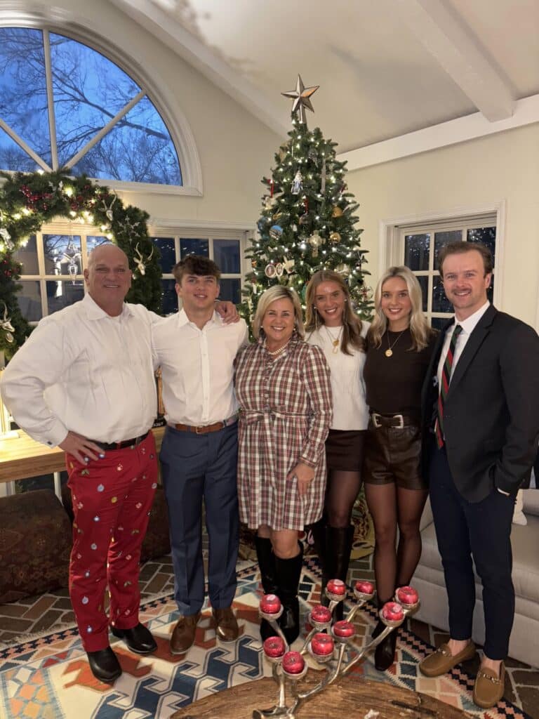 A family of six poses and smiles in front of a decorated Christmas tree inside a festive living room.