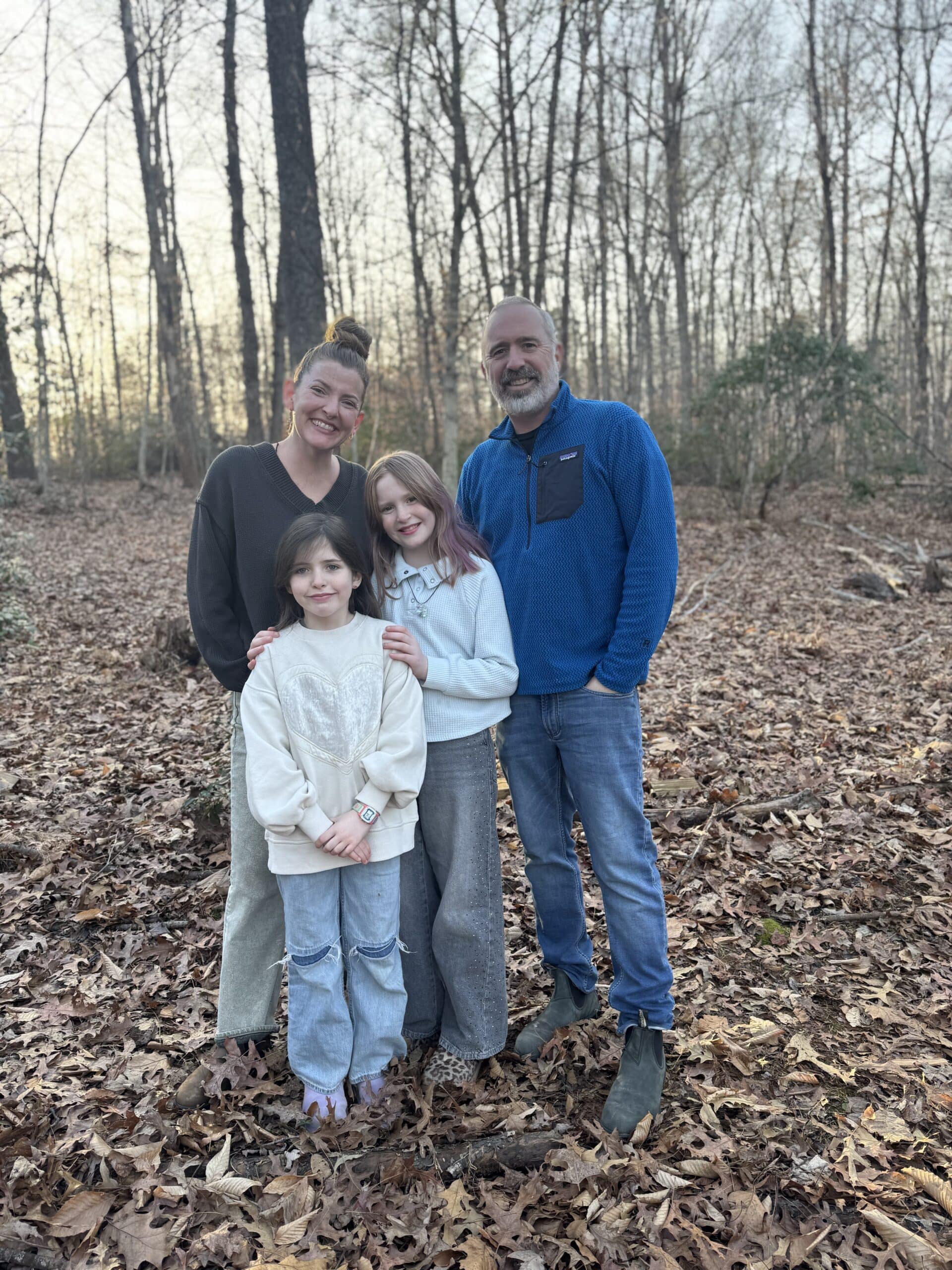 A family of four poses together in a forest with fallen leaves covering the ground.