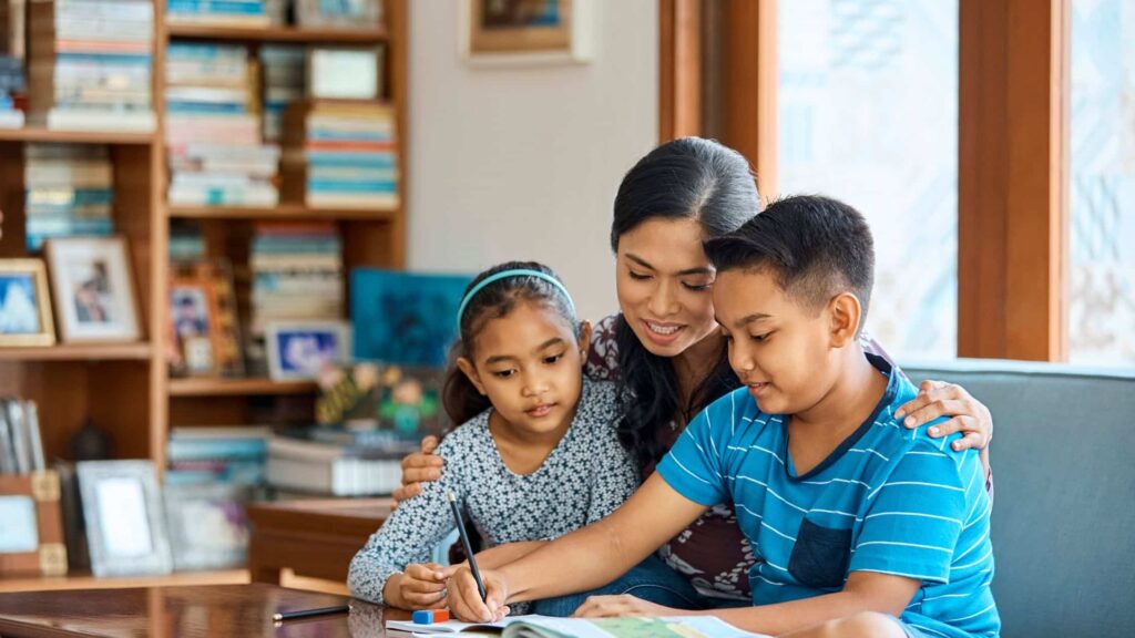 A woman helps two children with homework at a table in a cozy, book-filled room.