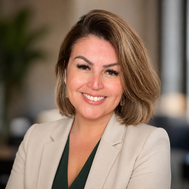 Smiling woman with short brown hair in a beige blazer, standing indoors with a blurred background.