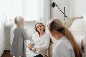 A woman sits on the floor talking to a child and another woman in a bright, cozy room.