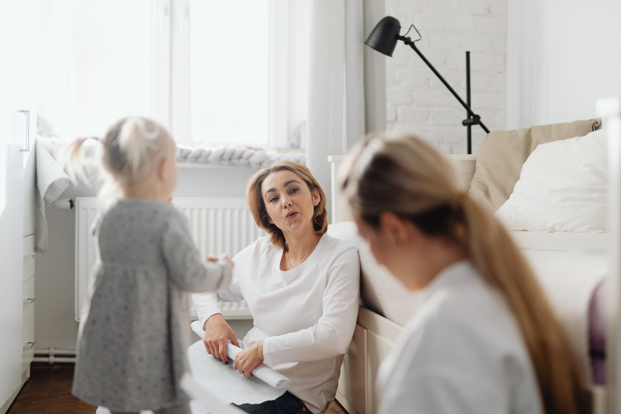 A woman sits on the floor talking to a child and another woman in a bright, cozy room.
