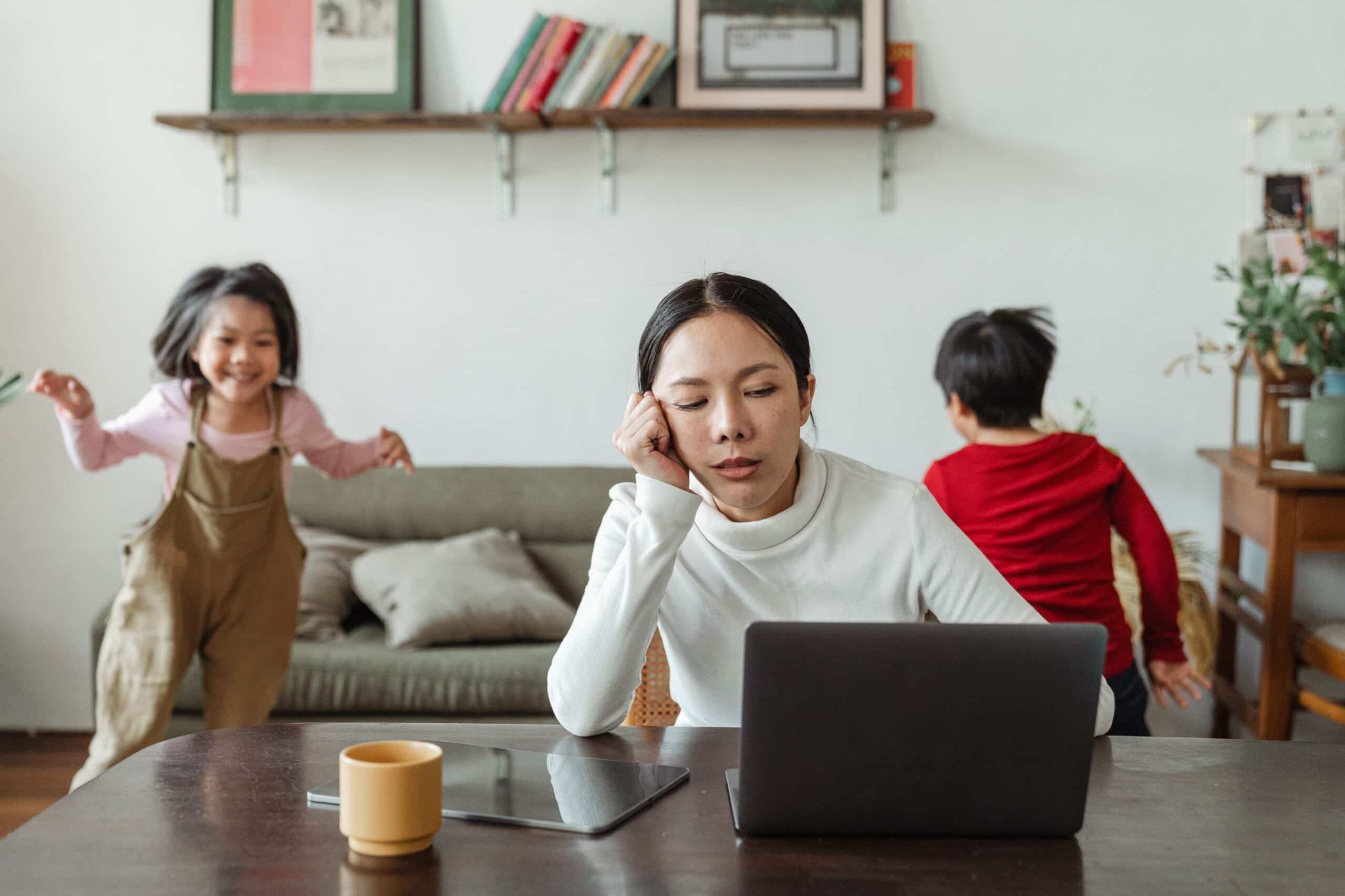A tired woman looks at a laptop while two children play in the background.