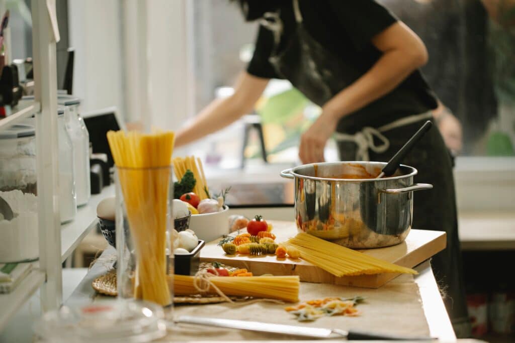 A person in a kitchen preparing a meal with pots, pasta, and fresh ingredients on the counter.